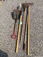 Five garden tools laid out on gravel - two shovels, a rake, a hoe, and one more shovel-like tool, showing wear and some rust on metal parts