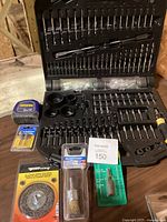 Photo showing open black plastic case with varied metal drill bits including spade bits, screwdriver bits, and other accessories, plus additional wire brushes and tape measure visible around the case.