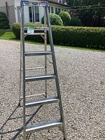 Side view of the 5ft aluminum step ladder standing on gravel, showing multiple wide steps and silver aluminum frame with a blue top plastic platform.