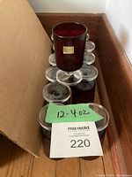 Top-down photo showing twelve red footed glasses stacked in two columns with clear protective covers, in a cardboard box with lot information visible.