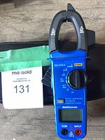 Front view of blue handheld digital clamp meter on carrying case with lot tag