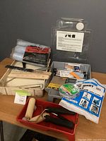 Multiple painting supplies arranged on a wooden table with trays, roller covers packet, paint rollers, brushes, adhesive tape and a packaged drop sheet in front of a dark gray wall.