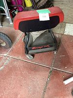 Rear view of the Snap-on mechanics creeper seat showing black metal frame with Snap-on logo on the tool tray and rear of red molded plastic seat.