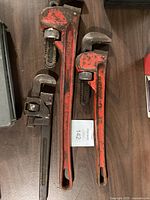 Three steel pipe wrenches of varying sizes (12", 18", 24") laid out on a dark surface, showing general wear and red paint chipping.