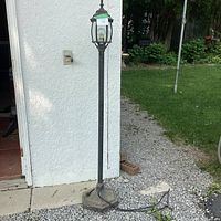Full view of the black metal outdoor lamp post standing next to a white wall and gravel ground showing the entire post and power cord.