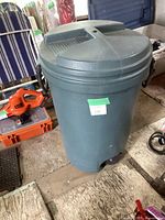 Photo showing green wheeled trash bin with closed round lid and side handle, positioned on wooden floor with other items around.