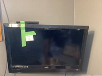 Vizio E320VL flat screen TV shown mounted on wall, screen black and off, green tape on bezel with sticker.