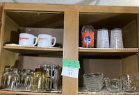 Photo showing upper and lower shelves with various mugs, glasses, and glass bowls arranged inside a wooden cabinet.