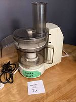 Full view of the food processor showing the transparent processing bowl, feed chute, lid, and cream colored base with a green label 'Tested + Works'