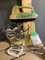 Yellow Iona R28B-1 stand mixer with glass bowl and metal beaters, cords coiled inside the bowl. Paper tag and tape marked 'Tested Works' attached to the unit.