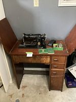 Wide angle photo showing the Singer sewing machine mounted on a wooden sewing table with drawers, foot pedal, manuals, and accessory box arranged on top.