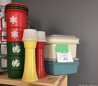 Four stacking mugs, two red featuring white snowflakes, two green featuring white roosters, each with lids, alongside yellow and red condiment containers, all displayed upright.