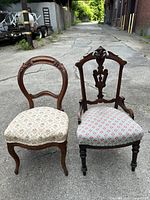 Two antique upholstered wooden side chairs placed side by side on a concrete surface outdoors. One chair has a rounded back and light floral fabric, the other has an ornate carved back with a female figure and pink patterned upholstery.