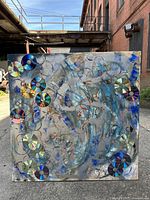 Overall front view of the square abstract mix-media painting with attached CDs outdoors under natural light on pavement against brick wall.