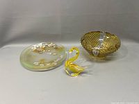 Full view of three art glass items on a light background: yellow glass swan, amber swirl-patterned bowl, and round foil fleck dish with floral design.
