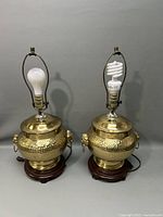 Pair of brass figural table lamps with different bulbs shown on top and polished wooden bases, against gray background.