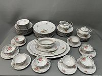 Full set arranged showing plates, bowls, cups with saucers, platters, creamer, sugar bowl and gravy boat with floral rose pattern and gold trim.