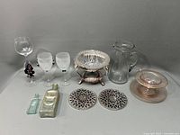 Wide view of all items including stem glasses, vintage medicine bottles, silverplate pedestal bowl, glass jug, metal trivets, and pink glass plates.