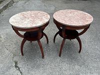Pair of round end tables with pink marble tops and wooden spider legs from front angle outside on pavement.