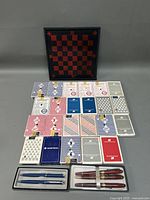 The lot arranged with 19 decks of vintage playing cards displayed in front of a red and black checkerboard, with two sets of vintage writing pens in cases at the front bottom.