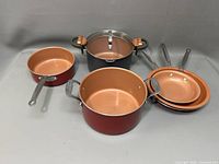 Cookware set including two red copper saucepans, one large black Gotham pot with copper interior and lid, and two red copper frying pans.