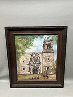 Framed oil painting showing Spanish mission-style church with people and horse at base, set against leafy background and sky.