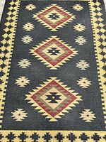 Close-up photo of the Kilim area rug showing the geometric diamond pattern in red, cream, black, and gold on a dark background.