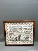 Full view of framed needlepoint sampler with alphabets and folk art scene, displayed against a gray background.