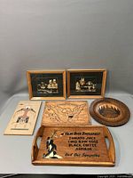 Photo showing the full set of 6 wooden decorative pieces arranged on a grey backdrop including framed art pieces, carved plate, tray with text, plaque and sand art panel.