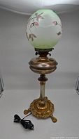 Full lamp showing ornate claw foot base, clear glass column, and frosted green hand-painted floral glass globe with cord.