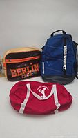 Three pre-owned bags shown on a white surface: one black-orange Berlin bag, one blue Dominaru backpack, and one red Cheer America duffel bag.
