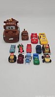 Front view of all 14 toy cars arranged on a plain background, showing different sizes and colors, including Mater, Lightning McQueen, Dinoco, and other vehicles from Disney Pixar Cars movie.