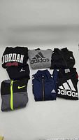 Seven folded kids sweaters and a pant set laid out showing Adidas and Nike logos in different colors and styles.