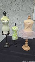 The set of three medium miniature dress forms shown on a black cloth background with a stucco wall behind. Includes one cream-colored fabric form with black and brown trim on a dark wood stand, one small green and white leaf-patterned form on a black metal stand, and a beige fabric form with French text and pink tulle tutu on a light wood stand.