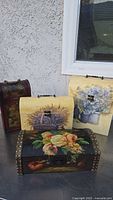 Four wooden decorative boxes shown on an outdoor surface against a wall and window background. Boxes vary in size and feature distinct floral decorative prints and bronze accent details.