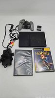 Overview of the PlayStation 2 Slim console, silver controller, memory card, power adapter, AV cable, and two game cases (The Hobbit and NCAA 2005 Football). All organized flat on a white surface.