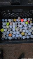 Top-down view of crate with assortment of 85 pre-owned golf balls of various brands in multiple colors including white, yellow, orange, pink, and neon, showing signs of wear and dirt.