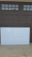 Large whiteboard with silver frame, clean surface, placed standing against a dark garage door outdoors.