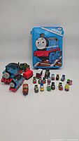 Various Thomas and Friends train toys displayed with blue storage case featuring Thomas on lid.