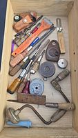 Top view of the drawer containing various vintage hand tools such as saw, screwdrivers, augers, measuring tapes, oil can, and leather sheathes.