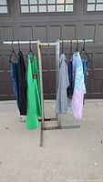 Five women's clothing items hanging on a metal stand outdoors against a garage door, items include dresses, tops, and a jacket in various colors and patterns.