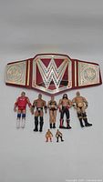 Photo showing 8 WWE wrestling action figures in front of a WWE Universal Champion Belt with sound feature, arranged on a grey background.