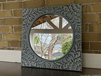 Front view of square mirror with round reflective surface and embossed tin frame with detailed floral pattern.