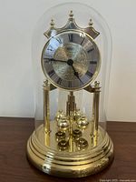 Full view of the Birks anniversary clock under a clear glass cloche showing the clock face, brass tone pillars, and rotating pendulum balls on the brass tone base.
