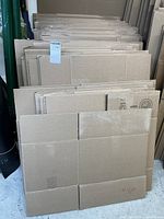 Flat-packed stacks of medium and large cardboard moving boxes standing upright against a wall, showing multiple layers and sizes of boxes in brown corrugated cardboard.