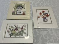 Three English floral watercolor prints showing varied flower and garden themes, signed, on white mats and laid on a fabric surface.