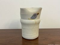 White porcelain vase with irregular four-lobed rim, hand-painted leaf or floral design near top, slight marks on surface, sitting on wooden surface against white background.