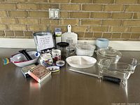Overview of kitchenware and decor items on a metal surface against a brick wall, showing glass dishes, ceramic bowls, a black-handled utensil, metal bucket, and other small items.
