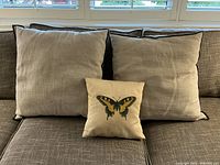 Three pillows on grey sofa: two large linen pillows with dark piping behind a smaller cream butterfly print pillow