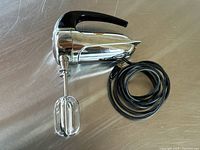 Side view of the Sunbeam Mixmaster Junior handheld mixer showing chrome finish, black handle, red power button, attached power cord, and one beater attached.